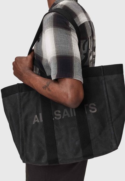 Personne portant une chemise à carreaux noire et blanche et portant un grand sac cabas noir avec "ALLSAINTS" imprimé sur le côté.