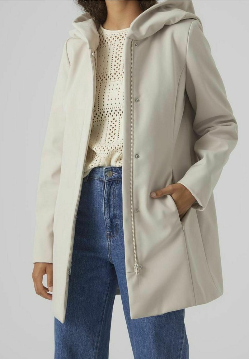 Veste à capuche beige clair en matière lisse, dotée d'une fermeture éclair à l'avant, de pressions et de poches latérales. Portée par-dessus un haut en maille.