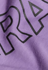 Fioletowy materiał o teksturowanej powierzchni, z wyraźnym czarnym napisem częściowo zakrytym, wskazującym na odzież codzienną.
