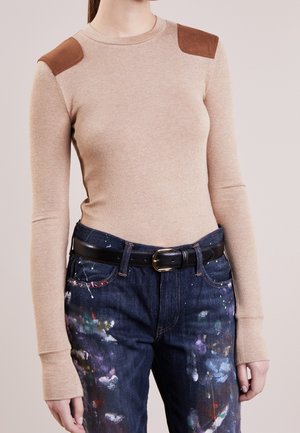 Beige trui met lange mouwen en bruine schouderstukken, gecombineerd met donkerblauwe spijkerbroek met meerkleurige verfvlekken en een zwarte riem.