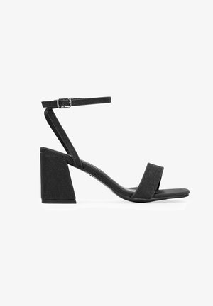 Black heeled sandals feature a textured fabric upper, an adjustable ankle strap with a silver buckle, and a chunky block heel.