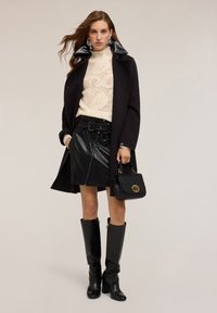 Abrigo negro con cuello brillante, suéter de punto crema, falda de cuero negra con cremallera, botas negras hasta la rodilla y bolso negro pequeño con acento dorado.