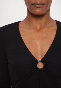 Femme portant un haut noir à col en V avec un détail d'anneau circulaire au centre, montrant le haut de la poitrine et le bas du visage.