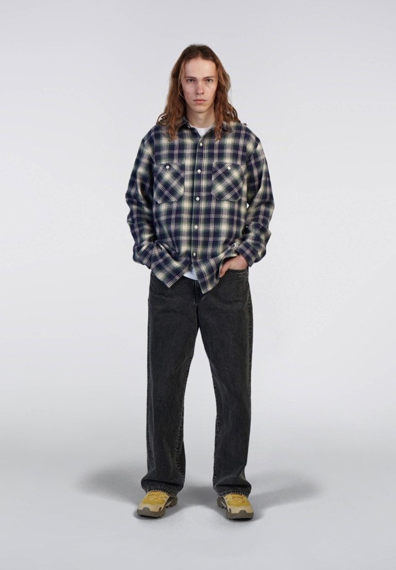 Camicia in flanella a quadri blu navy e verde, con due tasche sul petto. Abbinata a jeans baggy grigi scuri e sneakers gialle. Sfondo neutro.