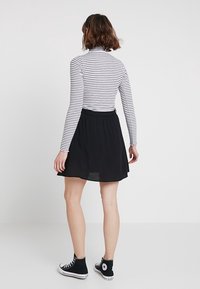 Kvinna med kort brunt hår iförd en svart kjol, svarta högklackade sneakers och en långärmad vit och grårandig topp, stående mot en vit bakgrund.