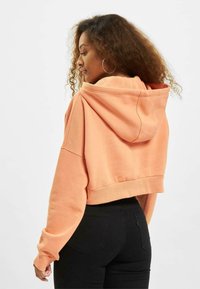 Sudadera recortada en naranja pastel, hecha de una tela suave, con capucha y mangas amplias, combinada con jeans negros ajustados.