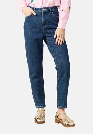 Mujer con vaqueros de tiro alto azules, camiseta rosa con detalles bordados y sandalias de cuero trenzado dorado, de pie con una mano en el bolsillo.