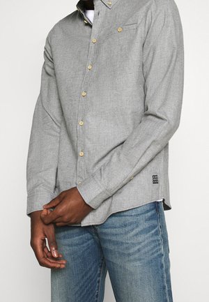 Homme portant une chemise gris clair à boutons jaunes et un jean bleu, mains jointes devant le corps.