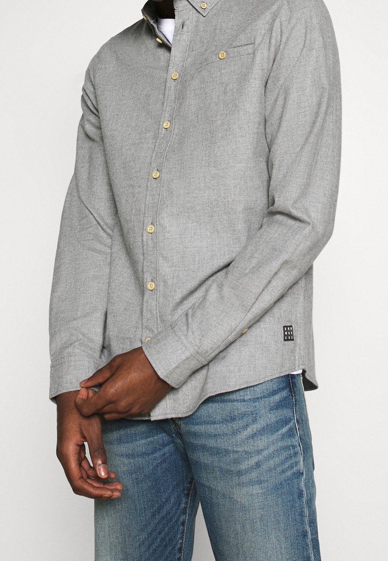 Homme portant une chemise gris clair à boutons jaunes et un jean bleu, mains jointes devant le corps.