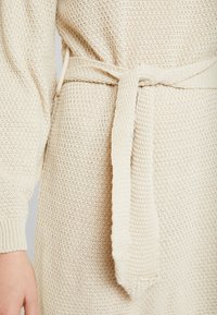 Robe en maille couleur crème avec manches longues et ceinture nouée à la taille. Présente un motif texturé et une coupe légèrement ample.