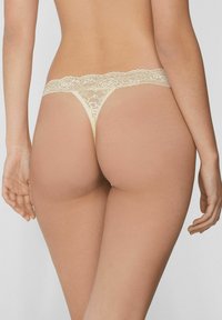 Beiger Spitzen-String mit dünnen Bändern und floralen Spitzen-Details, die ein freches Rücken-Design zeigen. Glatte Textur ergänzt zarte Akzente.