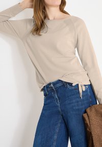 Femme portant un haut beige à manches longues et un jean bleu, tenant une veste marron texturée, posant devant un fond blanc.