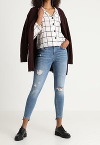 Camicia bianca a scacchi con bottoni neri, cardigan bordeaux, jeans skinny blu strappati e scarpe stringate nere.