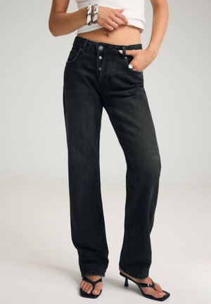 Femme portant un jean noir taille haute à jambes droites avec fermeture à boutons, un top court blanc, des bracelets argentés et des sandales noires à talons.