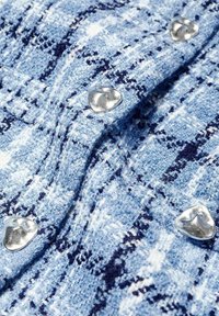 Lähikuva sinivalkoisesta teksturoidusta tweed-kankaasta, jossa epäsäännölliset hopeiset napit ruudukkomallissa.