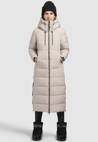Beige dlouhý péřový kabát s vysokým límcem, zapínáním na zip a elastickými akcenty. Texturovaný povrch s viditelnými horizontálními švy a bočními štěrbinami.