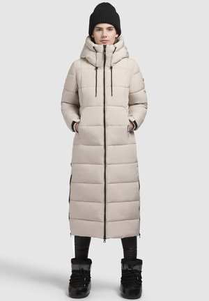 Longo cappotto imbottito beige con colletto alto, chiusura con zip e zip laterali. Presenta un motivo trapuntato e viene indossato sopra pantaloni neri e stivali.