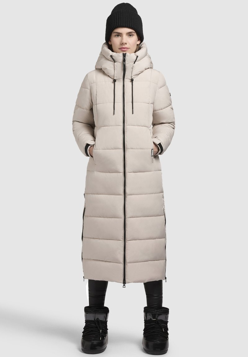 Beige dlouhý péřový kabát s vysokým límcem, zapínáním na zip a elastickými akcenty. Texturovaný povrch s viditelnými horizontálními švy a bočními štěrbinami.