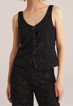 Kvinde iført en ærmeløs sort tekstureret vest med tre knapper og matchende højtaljede bukser mod en neutral baggrund.