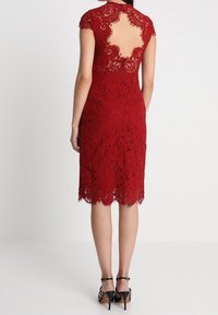 Femme portant une robe en dentelle rouge longueur genou avec une grande découpe en dentelle dans le dos, associée à des sandales à talons hauts noires.