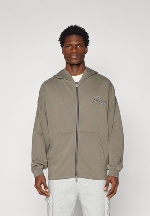 Hoodie gris zippé avec capuche, présentant un logo sur le devant, une poche kangourou et des poignets élastiques. Le tissu a une finition douce et texturée.