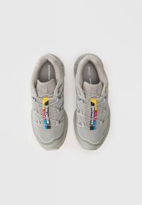 Ténis atléticos cinzas com material em malha, design em camadas, atacadores elásticos e um logótipo de marca em destaque. Apresenta uma aba multicolorida na lateral.