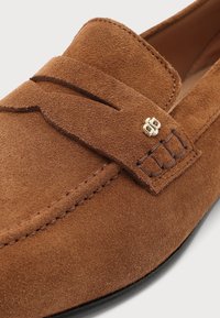 Gros plan sur un mocassin en daim marron avec une couture détaillée, une bride en cuir sur le dessus et un petit logo métallique doré sur le côté.