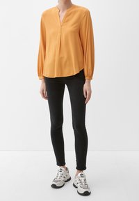 Blouse orange à col en V, manches longues et ourlet légèrement arrondi, associée à un jean skinny noir et des baskets blanches.