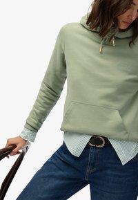 Sudadera verde claro con cordón, adornos dorados y un bolsillo frontal, superpuesta sobre una camisa a rayas azul y blanca, combinada con vaqueros azules.