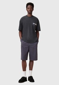 Sort bomulds T-shirt med logo-detalje på brystet, parret med knæ-lange blå og brune ternede shorts. Modellen bærer hvide ankelstrømper og sorte sko.