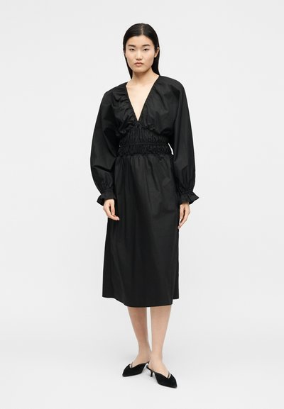 Γυναίκα με ίσια σκούρα μαλλιά, φορώντας μαύρο midi φόρεμα με μακριά μανίκια, βαθιά V λαιμόκοψη και ραμμένη μέση, σε συνδυασμό με μαύρες μυτερές γόβες.