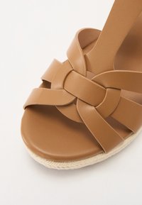Sandale en cuir marron avec des brides tressées, design à bout ouvert, et une semelle de style espadrille texturée. Finition lisse sans détails de quincaillerie visibles.