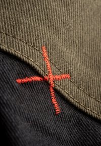 Tissu en coton de couleur vert olive et noir avec des coutures visibles. Présente un détail en point de croix rouge, mettant en valeur la couture entre les deux couleurs.