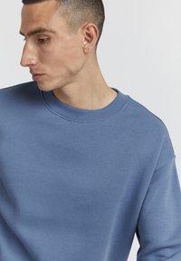 Kék pulóver kerek nyakkivágással, puha anyaggal és laza szabással, vállvarrással és sima felülettel.