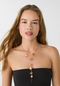 Giovane donna con capelli lunghi e ondulati, che indossa un top nero senza spalline e una collana d'oro con pendenti a forma di foglia, su uno sfondo bianco.