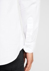 Camisa blanca de vestir con mangas largas, puños de botón y un dobladillo curvado. La tela parece suave y ligera, combinada con pantalones oscuros.
