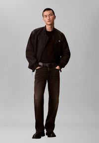 Jonge man met een zwart bomberjack, een zwart T-shirt, donkere spijkerbroek met een zwarte riem en zwarte schoenen, staand met handen in de zakken.