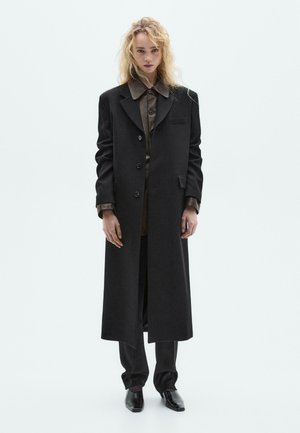 Long black coat with a notched collar, single-breasted design, front pockets, layered over a brown button-up shirt and dark trousers.