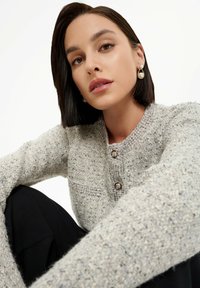 Szary sweter o teksturze z okrągłym dekoltem i dwoma dużymi guzikami, zestawiony z czarnymi spodniami. Eleganckie kolczyki w formie kropli podkreślają całość.