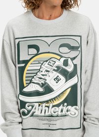 Persoon in een lichtgrijze sweatshirt met een grote afbeelding van een zwart-witte DC sneaker en de tekst "DC Athletics" aan de voorkant.
