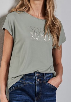 Mujer con camiseta verde claro con texto "SPARK FOUND" y vaqueros de doble botón azul oscuro, con una mano en el bolsillo, apoyada contra una pared.