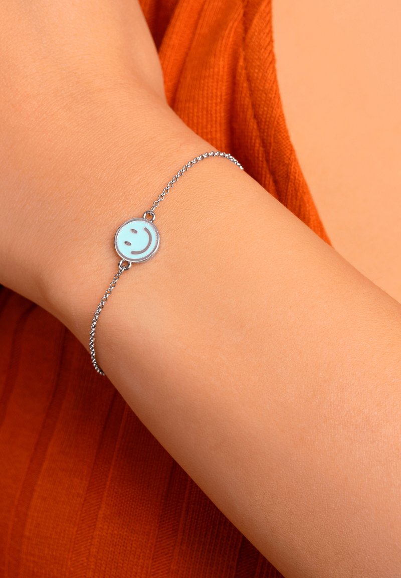 Silverfärgad kedjearmband med en rund, ljusblå berlock med ett leende ansiktsdesign. Ytan är slät, med en polerad finish.