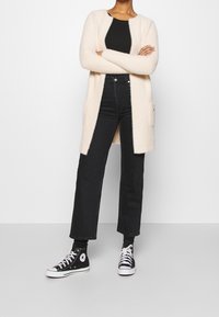 Dlouhý měkký krémový kardigan s otevřeným předním dílem, černé spodní tričko, černé džíny s vysokým pasem a černé tenisky Converse. Bez viditelných vzorů.