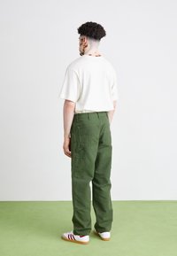 Groene cargobroek met meerdere zakken, gecombineerd met een beige t-shirt en witte sneakers met rode accenten. De achtergrond is een lichtgroen tapijt.