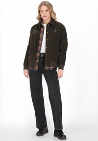 Młoda kobieta ubrana w czarną kurtkę z naszywką w kształcie kotwicy, w kratkowaną koszulę, czarne dżinsy i czarne botki, stojąca na białym tle.