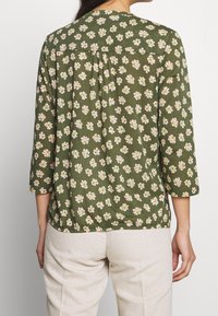 Vihreä kukkapaikka, jossa on takakiila, 3/4 hihaa ja lievästi kaareva helma. Pienet vaaleanpunaiset ja valkoiset kukat koristavat ympäriinsä.