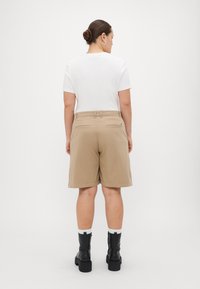 Khakifarbenen Shorts mit einem lockeren Schnitt, ausgestattet mit zwei Gesäßtaschen und einer glatten Textur, kombiniert mit einem weißen T-Shirt und schwarzen Stiefeletten.