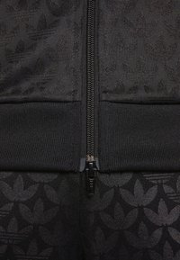 Svart zip-up jacka med ett strukturerat mönster av mörka blomformer. Har en kontrasterande slät dragkedja samt ribbade ärmslut och midja.