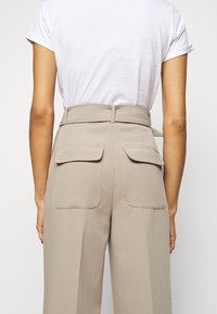 Pantalons beiges taille haute avec deux poches arrière, passants de ceinture latéraux et une texture lisse, associés à un t-shirt blanc.