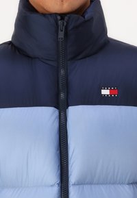 Tamno plavi i svijetloplavi pufer jakna s visokim ovratnikom, prednjim zipom i izvezenim logom "Tommy Jeans" na lijevoj strani prsnog dijela. Glatka, izolirana tekstura.